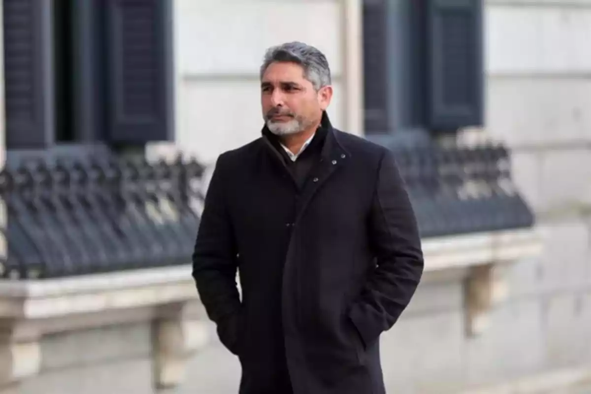 Hombre de cabello canoso y barba con abrigo oscuro caminando por la calle frente a un edificio antiguo Hombre de cabello canoso y barba con abrigo oscuro caminando por la calle frente a un edificio antiguo