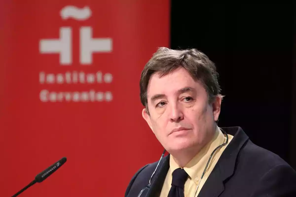 Hombre de cabello corto y canoso con traje y corbata, sentado frente a un micrófono con un fondo rojo que tiene el logotipo y el nombre del Instituto Cervantes desenfocados. Hombre de cabello corto y canoso con traje y corbata, sentado frente a un micrófono con un fondo rojo que tiene el logotipo y el nombre del Instituto Cervantes desenfocados.