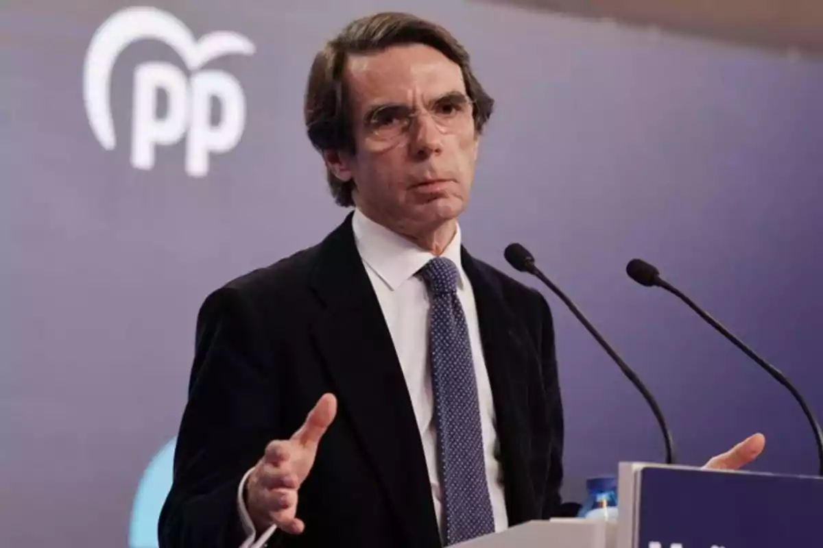 Hombre de cabello castaño y gafas hablando en un podio con dos micrófonos y el logotipo del PP en el fondo
