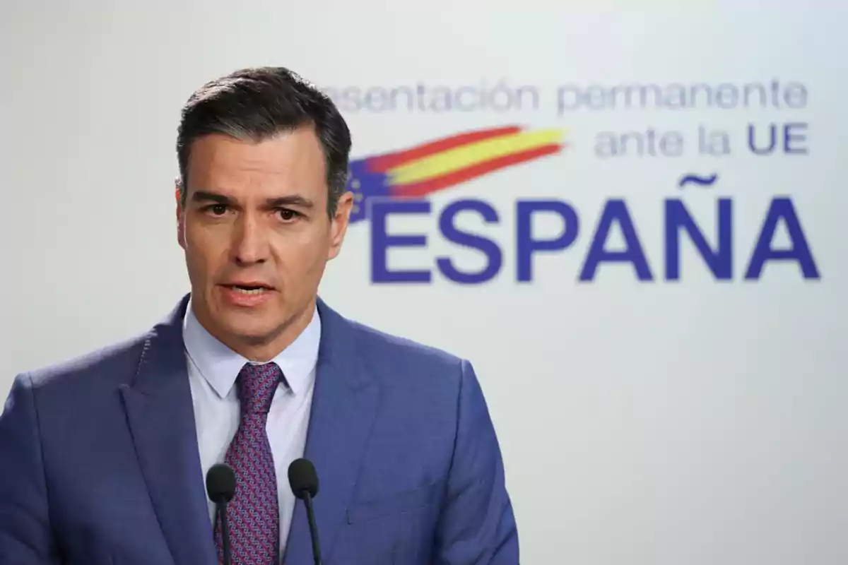 Hombre con traje azul hablando frente a micrófonos con un fondo que dice España y muestra una bandera estilizada Hombre con traje azul hablando frente a micrófonos con un fondo que dice España y muestra una bandera estilizada