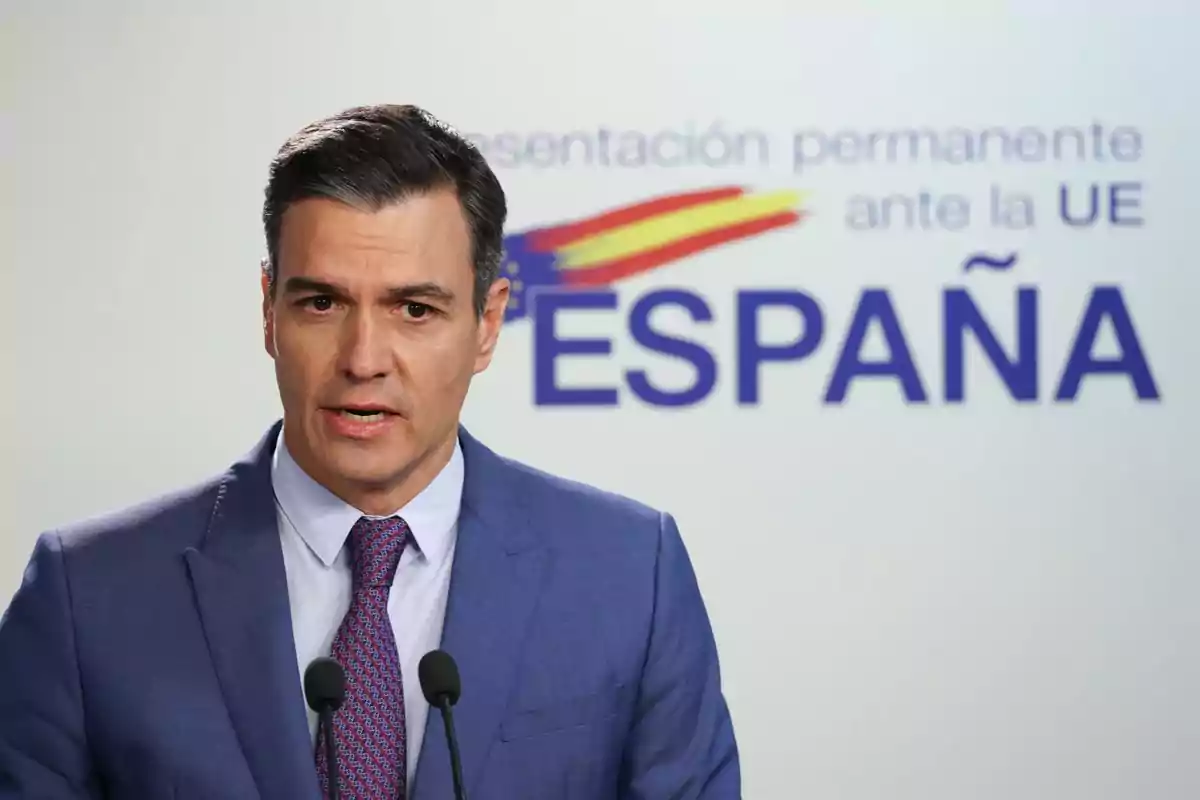 Man in a blue suit speaking in front of microphones with a Spain sign and the flag in the background