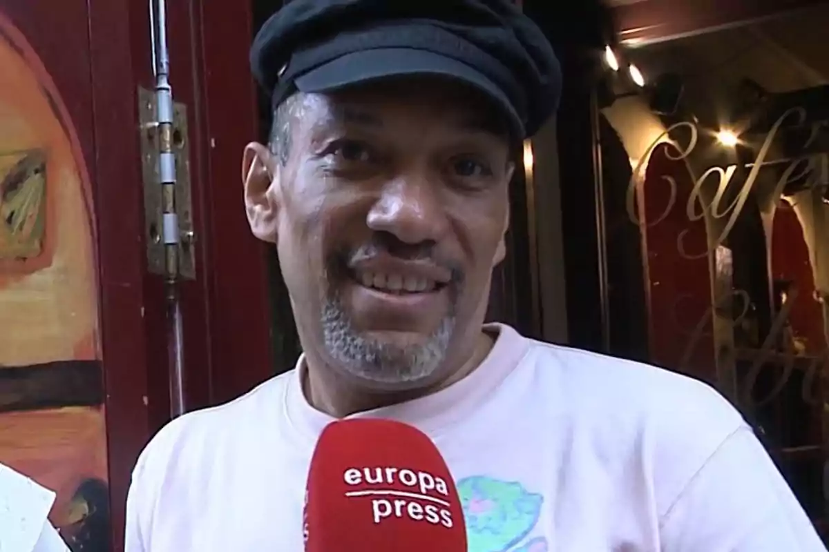 Hombre con gorra negra y camiseta clara sonriendo mientras sostiene un micrófono rojo de europa press frente a una cafetería Hombre con gorra negra y camiseta clara sonriendo mientras sostiene un micrófono rojo de europa press frente a una cafetería