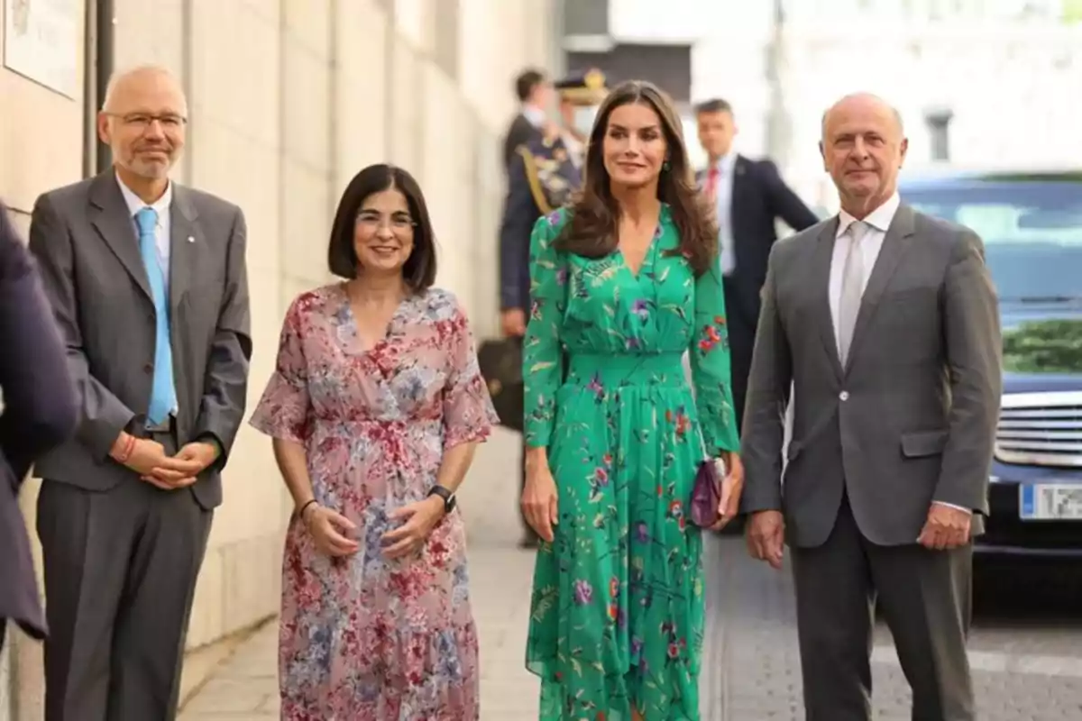 Cuatro personas posan sonrientes en la calle junto a un edificio, dos hombres con traje y corbata y dos mujeres con vestidos estampados, una de ellas con un vestido verde y la otra con un vestido rosa con flores