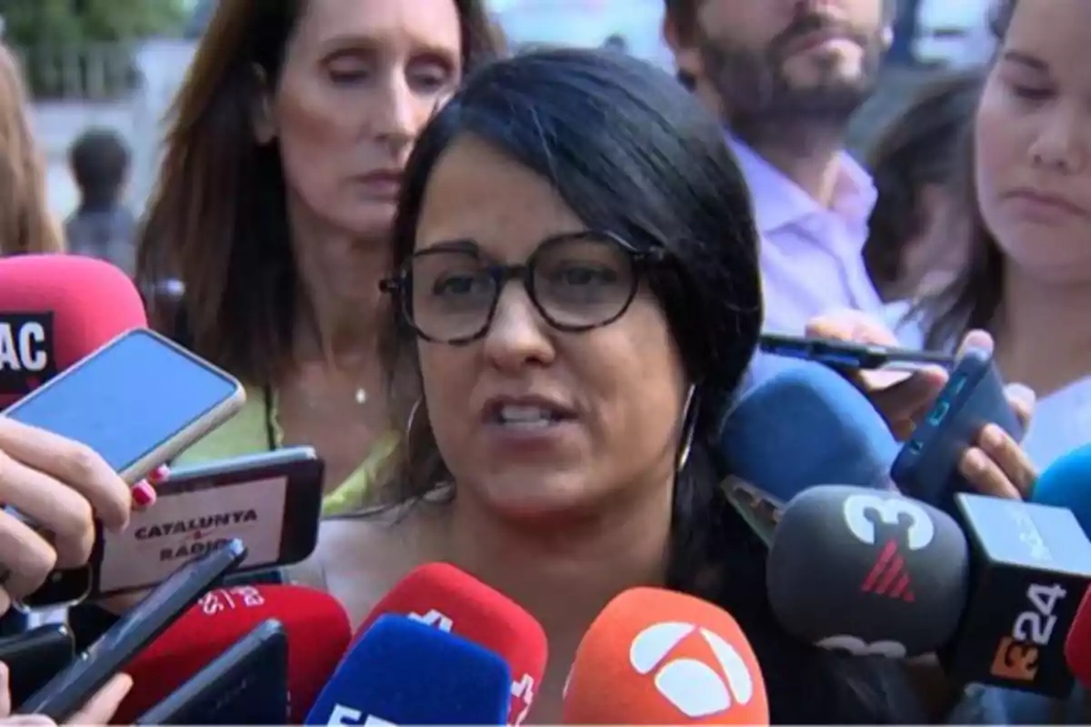 Mujer de cabello oscuro y gafas hablando con varios micrófonos de diferentes medios de comunicación frente a ella rodeada de periodistas Mujer de cabello oscuro y gafas hablando con varios micrófonos de diferentes medios de comunicación frente a ella rodeada de periodistas