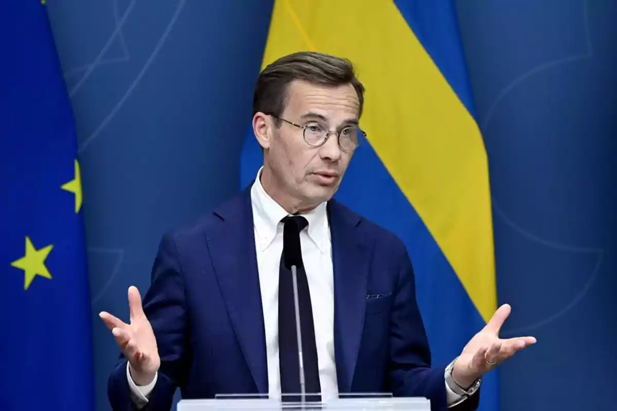 Hombre con gafas y traje azul hablando en un podio con banderas de la Unión Europea y Suecia de fondo Hombre con gafas y traje azul hablando en un podio con banderas de la Unión Europea y Suecia de fondo