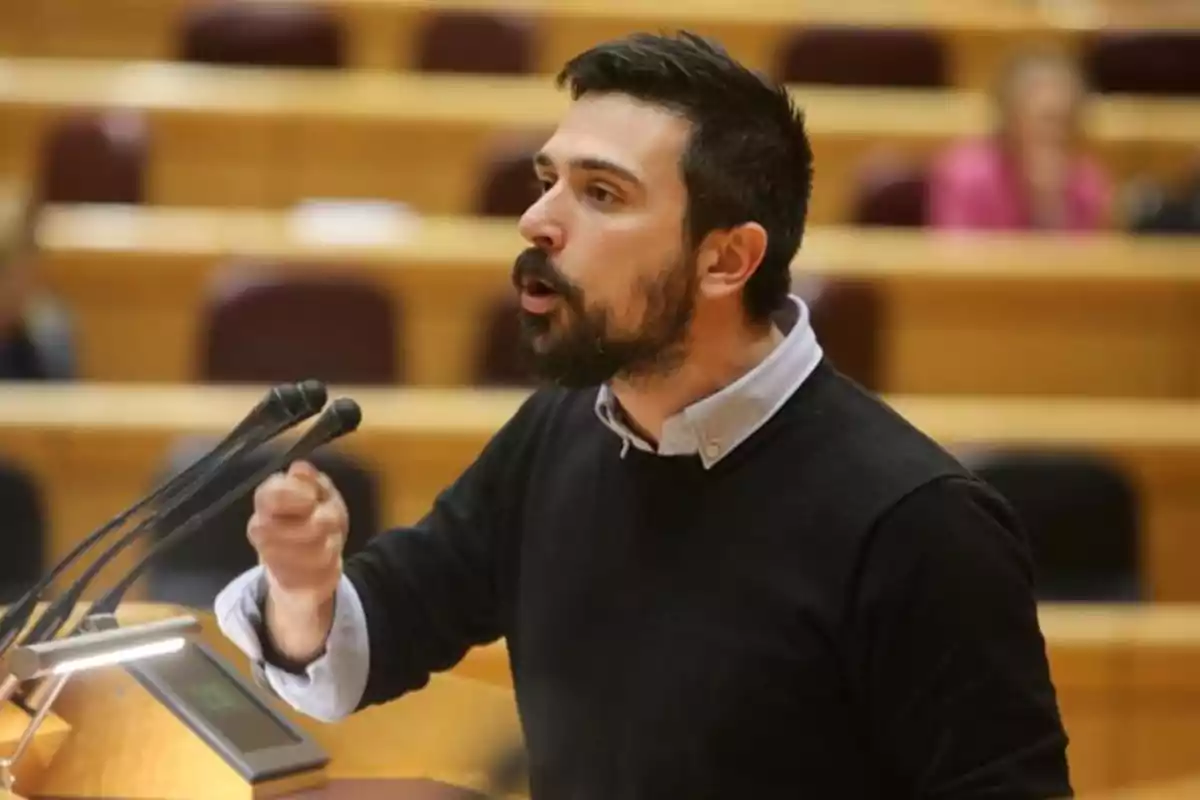 Hombre joven con barba hablando apasionadamente en un podio con micrófonos en un auditorio Hombre joven con barba hablando apasionadamente en un podio con micrófonos en un auditorio