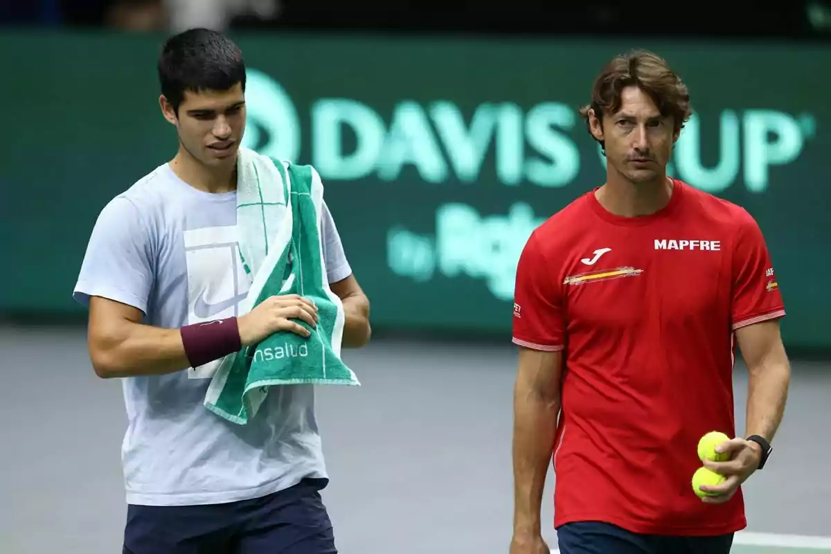 Dos hombres en una cancha de tenis uno con camiseta clara y toalla verde y el otro con camiseta roja sosteniendo pelotas de tenis Dos hombres en una cancha de tenis uno con camiseta clara y toalla verde y el otro con camiseta roja sosteniendo pelotas de tenis