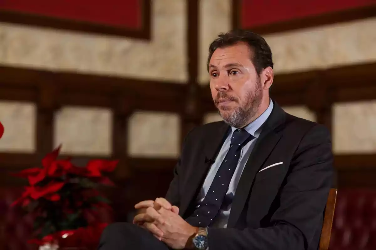 Hombre con barba y traje oscuro sentado en una silla con las manos entrelazadas en un ambiente elegante con una planta de flores rojas al fondo Hombre con barba y traje oscuro sentado en una silla con las manos entrelazadas en un ambiente elegante con una planta de flores rojas al fondo
