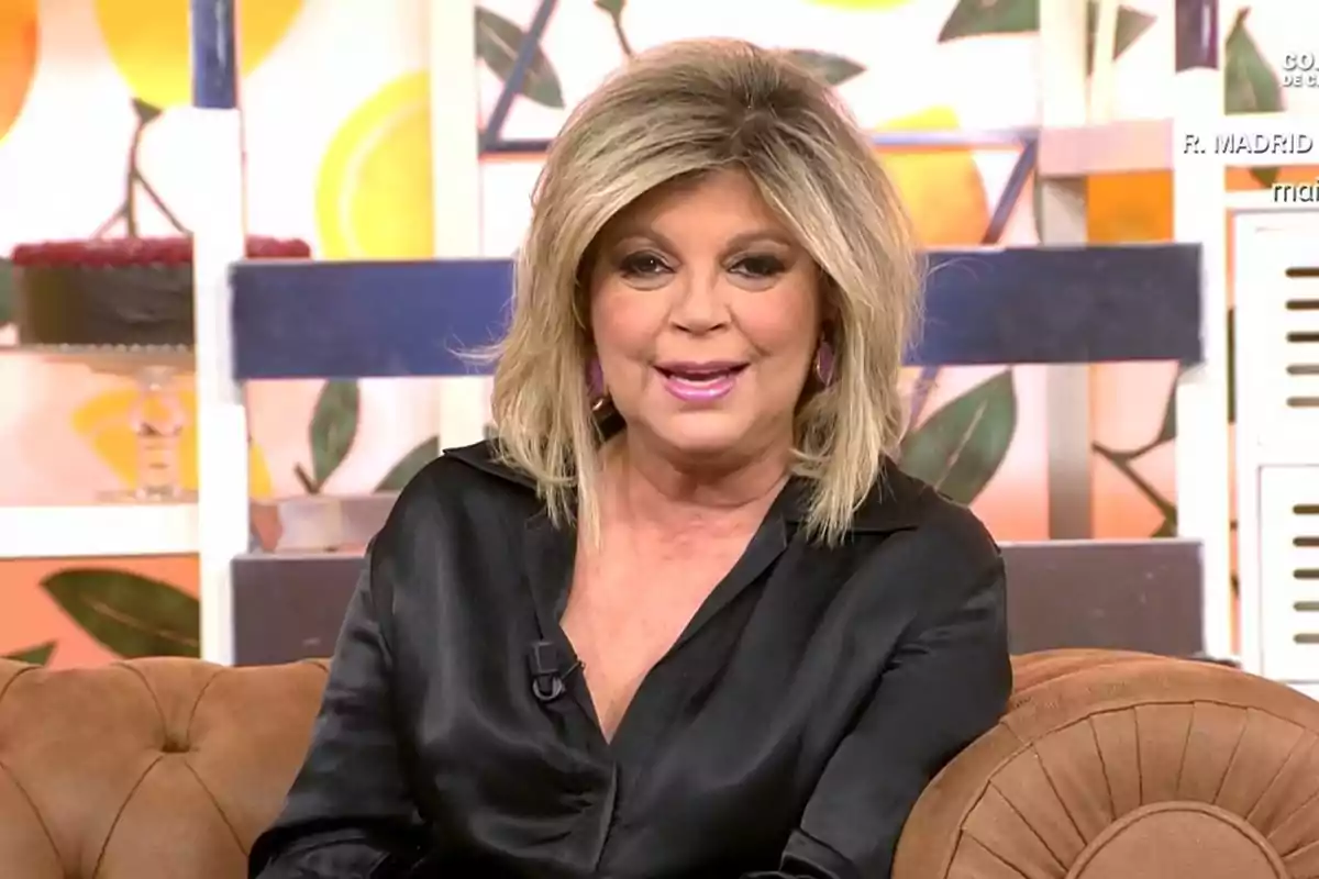 Mujer rubia con blusa negra sentada en un sofá marrón en un set de televisión colorido Mujer rubia con blusa negra sentada en un sofá marrón en un set de televisión colorido