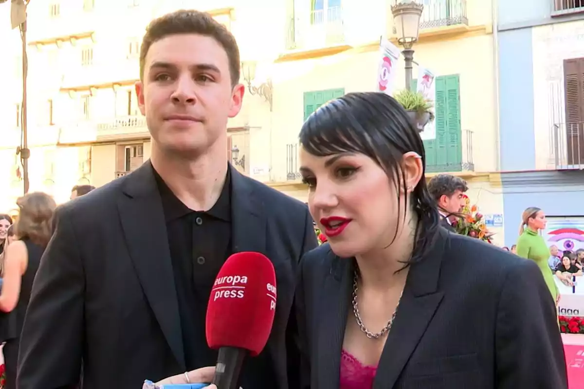 Dos personas vestidas de negro son entrevistadas en un evento al aire libre mientras sostienen un micrófono rojo de Europa Press Dos personas vestidas de negro son entrevistadas en un evento al aire libre mientras sostienen un micrófono rojo de Europa Press