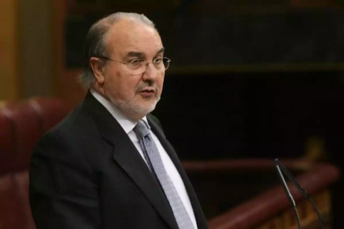 Hombre mayor con barba y gafas hablando en un podio dentro de un recinto parlamentario Hombre mayor con barba y gafas hablando en un podio dentro de un recinto parlamentario