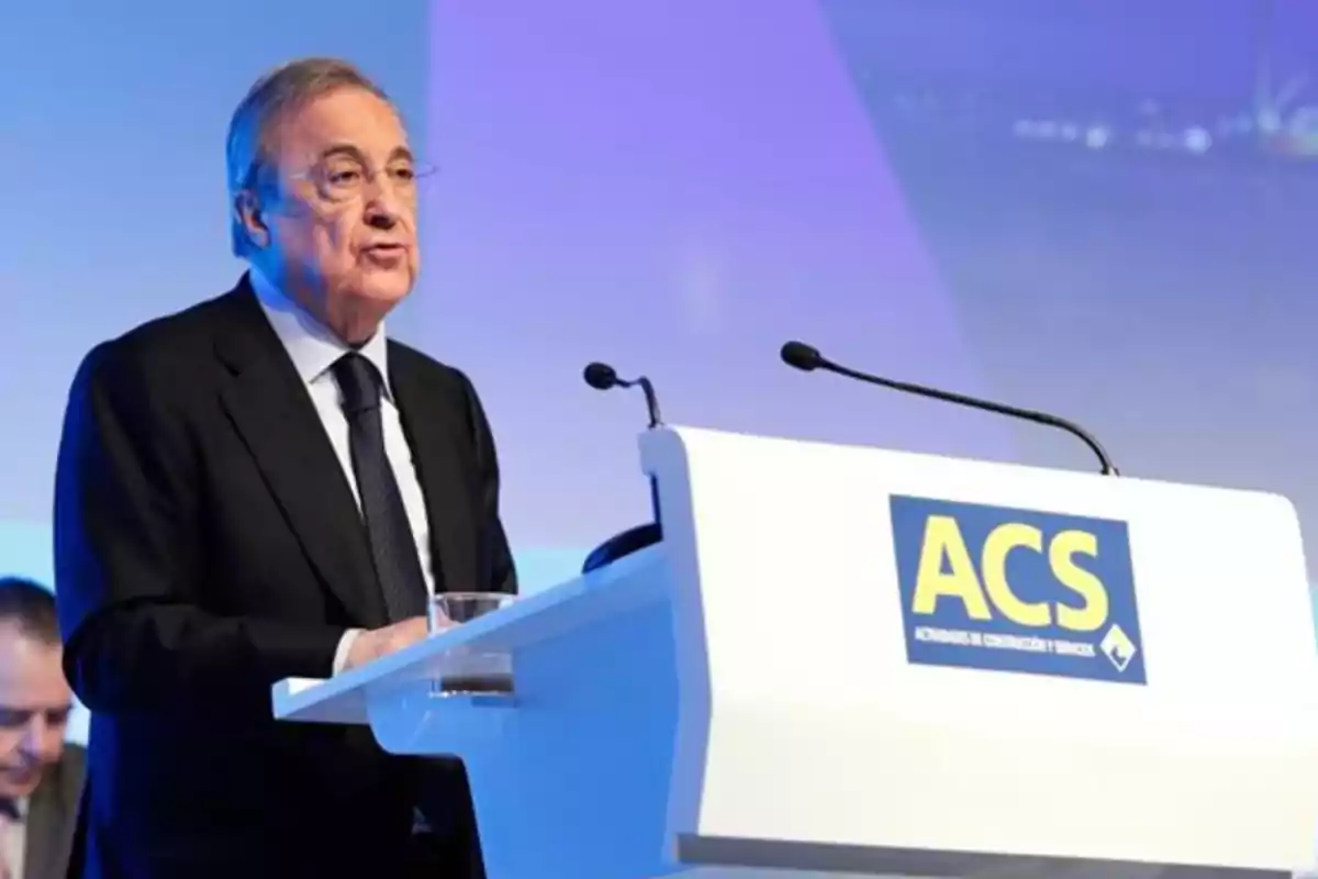 Hombre de cabello canoso con gafas y traje oscuro hablando en un podio blanco con el logo de ACS en un evento empresarial Hombre de cabello canoso con gafas y traje oscuro hablando en un podio blanco con el logo de ACS en un evento empresarial