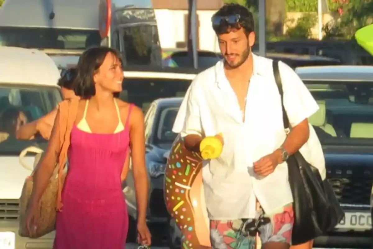 Una pareja joven camina por la calle en un día soleado, ella lleva un vestido rosa y un bolso de mimbre, él viste una camisa blanca y lleva un flotador de dona y una bolsa negra, ambos parecen contentos y relajados Una pareja joven camina por la calle en un día soleado, ella lleva un vestido rosa y un bolso de mimbre, él viste una camisa blanca y lleva un flotador de dona y una bolsa negra, ambos parecen contentos y relajados
