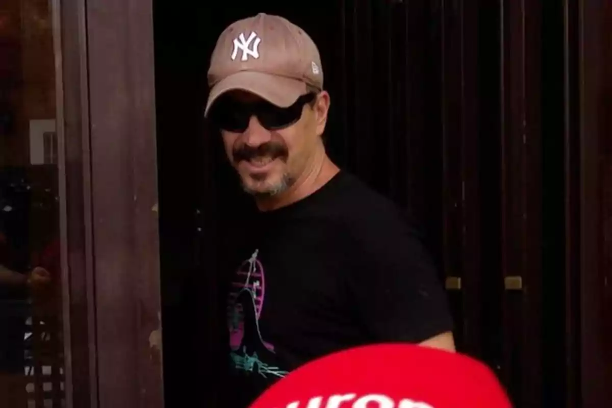 Hombre con gorra beige de los Yankees, gafas de sol y camiseta negra sonriendo cerca de una puerta Hombre con gorra beige de los Yankees, gafas de sol y camiseta negra sonriendo cerca de una puerta