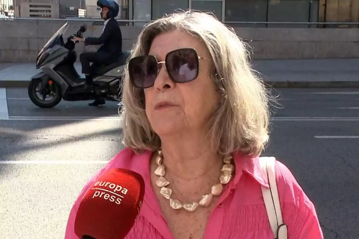 Mujer de cabello canoso y gafas grandes siendo entrevistada en la calle con un micrófono rojo de europa press mientras un hombre en moto pasa al fondo Mujer de cabello canoso y gafas grandes siendo entrevistada en la calle con un micrófono rojo de europa press mientras un hombre en moto pasa al fondo
