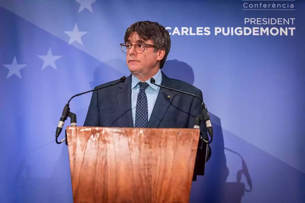 Hombre con gafas y traje azul hablando en un podio de madera frente a un fondo azul con estrellas y texto. Hombre con gafas y traje azul hablando en un podio de madera frente a un fondo azul con estrellas y texto.