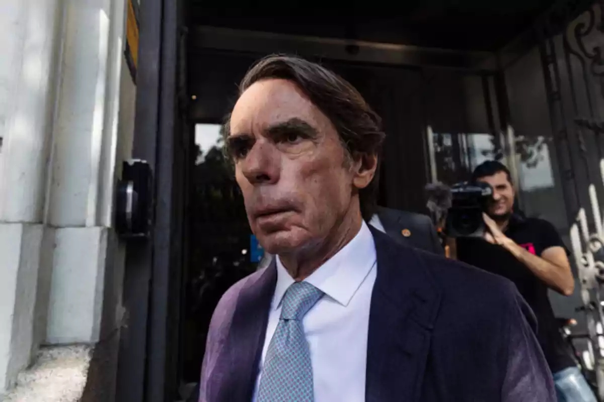 Hombre de cabello canoso y traje azul saliendo de un edificio mientras un camarógrafo lo graba al fondo Hombre de cabello canoso y traje azul saliendo de un edificio mientras un camarógrafo lo graba al fondo