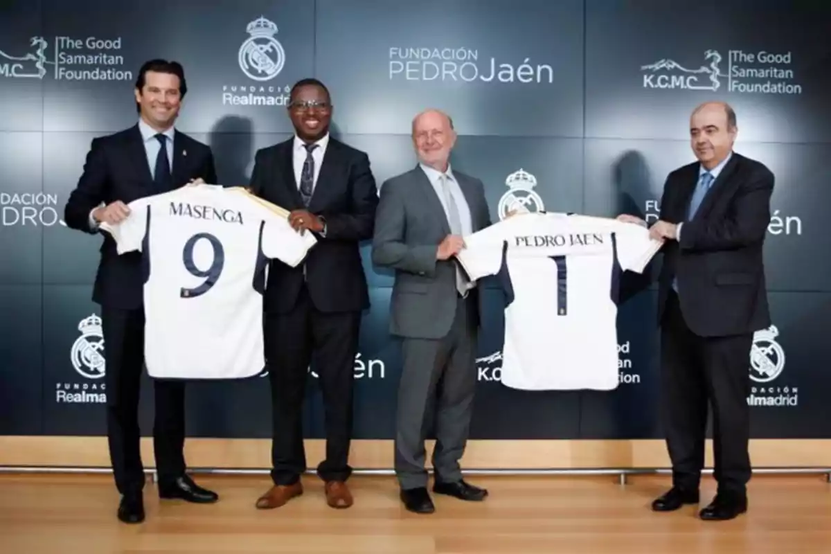 Cuatro hombres de traje posan sonrientes sosteniendo camisetas del Real Madrid personalizadas en un evento institucional con logotipos de la Fundación Real Madrid y otras organizaciones en el fondo Cuatro hombres de traje posan sonrientes sosteniendo camisetas del Real Madrid personalizadas en un evento institucional con logotipos de la Fundación Real Madrid y otras organizaciones en el fondo