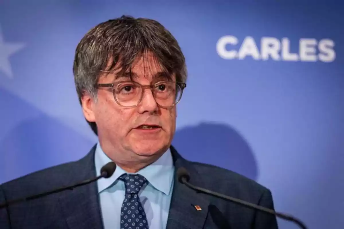 Hombre de cabello canoso y gafas hablando frente a micrófonos con fondo azul donde se lee la palabra CARLES