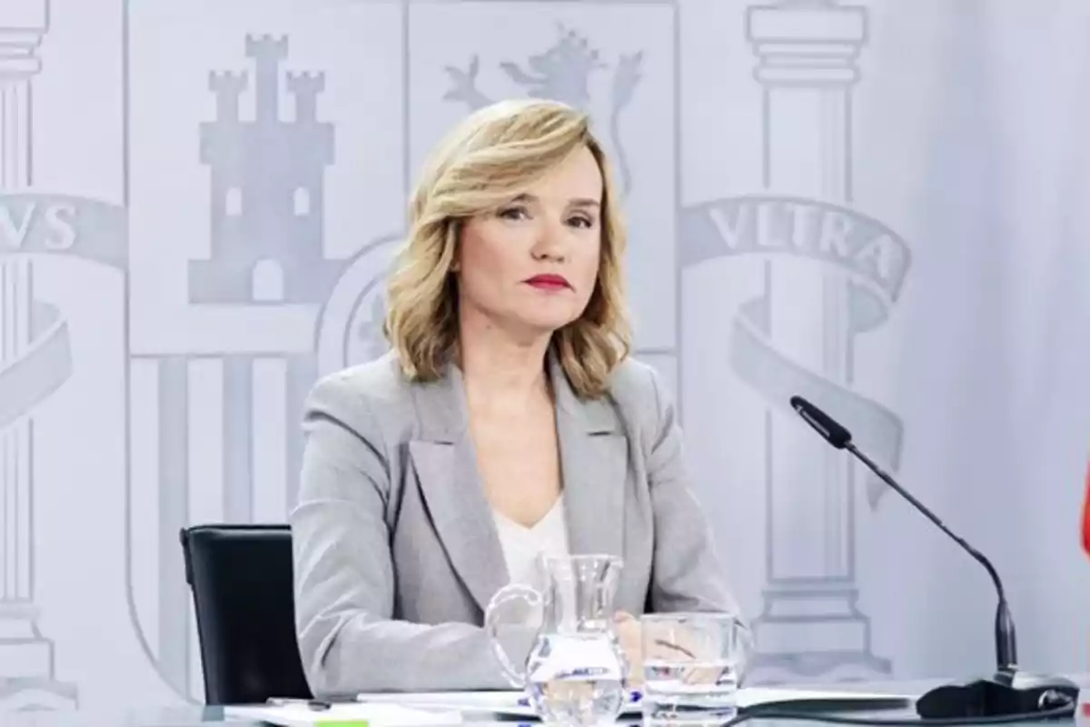 Mujer rubia con chaqueta gris sentada en una mesa con micrófono y fondo institucional