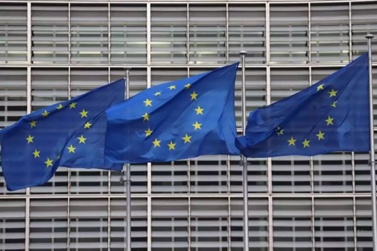 Tres banderas de la Unión Europea ondeando frente a un edificio con ventanas. Tres banderas de la Unión Europea ondeando frente a un edificio con ventanas.