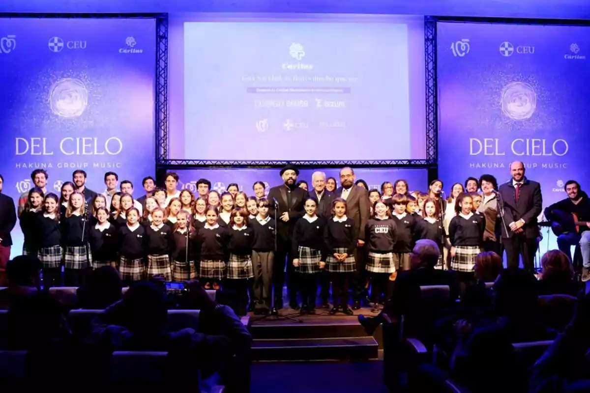 Un grupo numeroso de niños y adultos posa en el escenario de un auditorio durante un evento musical llamado Del Cielo, con un fondo azul y pantallas que muestran información sobre Cáritas y CEU, mientras el público observa desde sus asientos. Un grupo numeroso de niños y adultos posa en el escenario de un auditorio durante un evento musical llamado Del Cielo, con un fondo azul y pantallas que muestran información sobre Cáritas y CEU, mientras el público observa desde sus asientos.