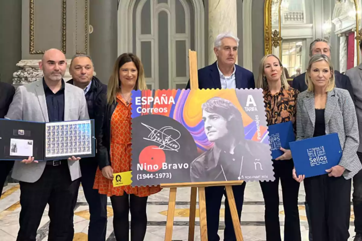 Un grupo de personas posa en un elegante salón junto a una imagen ampliada de un sello postal dedicado a Nino Bravo Un grupo de personas posa en un elegante salón junto a una imagen ampliada de un sello postal dedicado a Nino Bravo