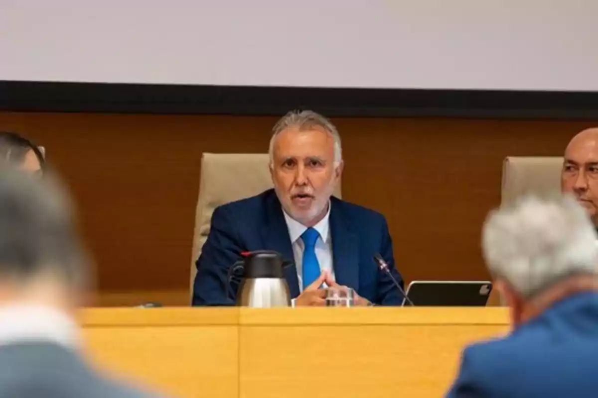 Gray-haired, bearded man sitting at a conference table in a blue suit and blue tie, speaking into a microphone in a meeting room Gray-haired, bearded man sitting at a conference table in a blue suit and blue tie, speaking into a microphone in a meeting room