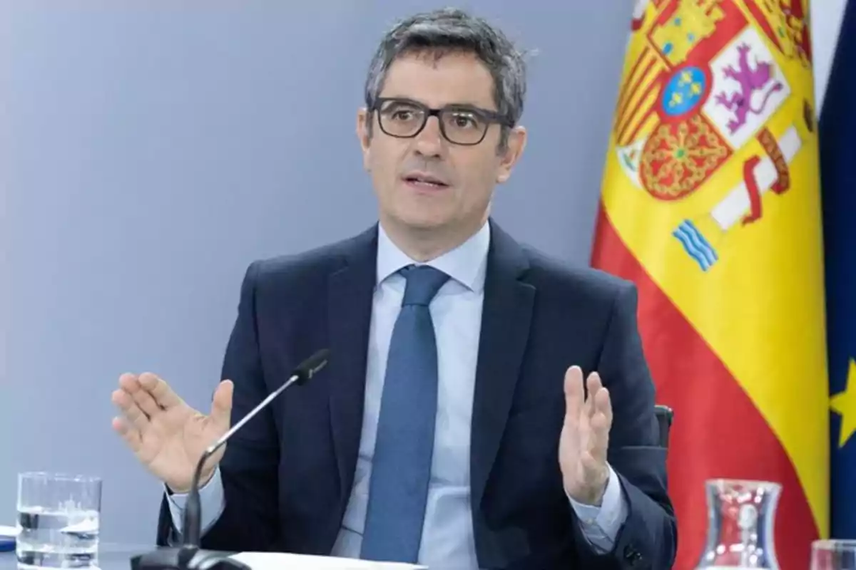 Hombre con gafas y traje azul hablando en una conferencia con una bandera de España al fondo Hombre con gafas y traje azul hablando en una conferencia con una bandera de España al fondo