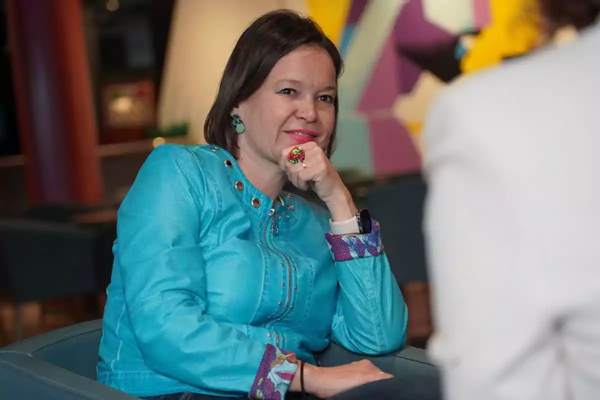 Mujer sentada con chaqueta azul turquesa y accesorios coloridos, apoyando el mentón en su mano y sonriendo durante una conversación en un ambiente interior colorido. Mujer sentada con chaqueta azul turquesa y accesorios coloridos, apoyando el mentón en su mano y sonriendo durante una conversación en un ambiente interior colorido.