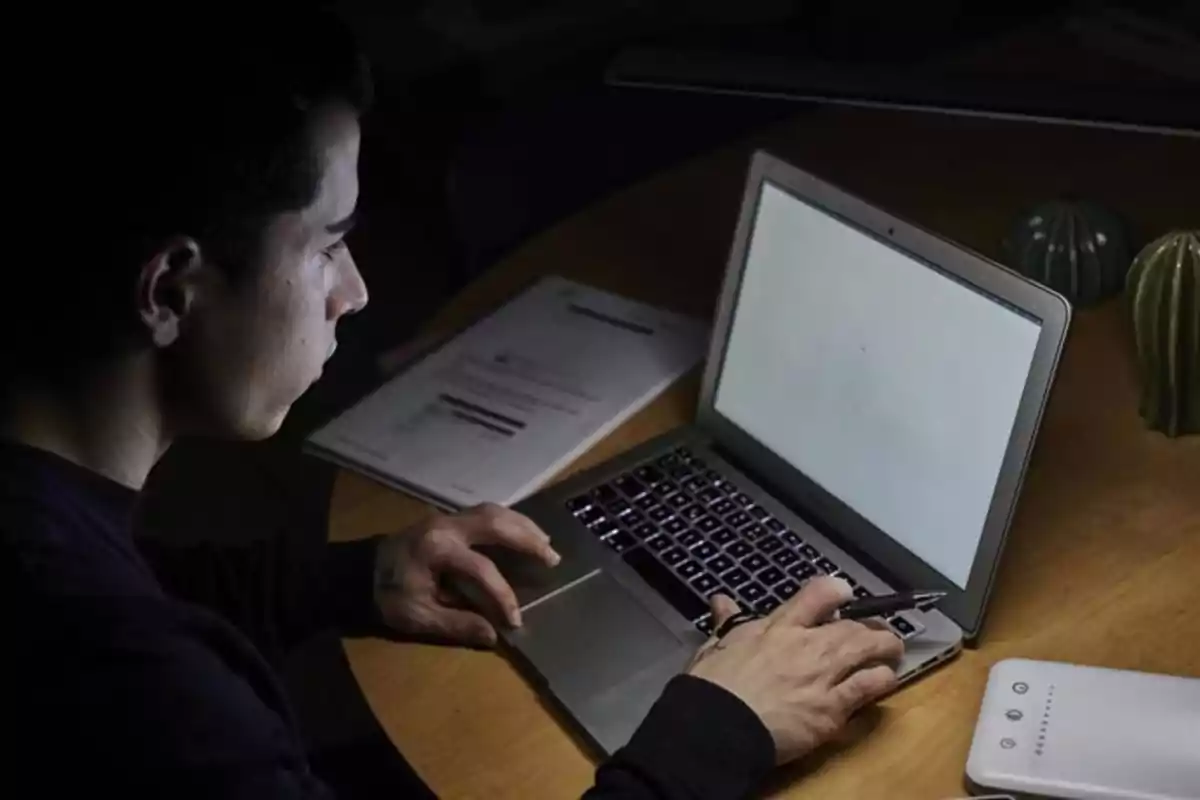 Persona usando una laptop con la pantalla en blanco mientras sostiene una pluma y tiene documentos sobre la mesa Persona usando una laptop con la pantalla en blanco mientras sostiene una pluma y tiene documentos sobre la mesa