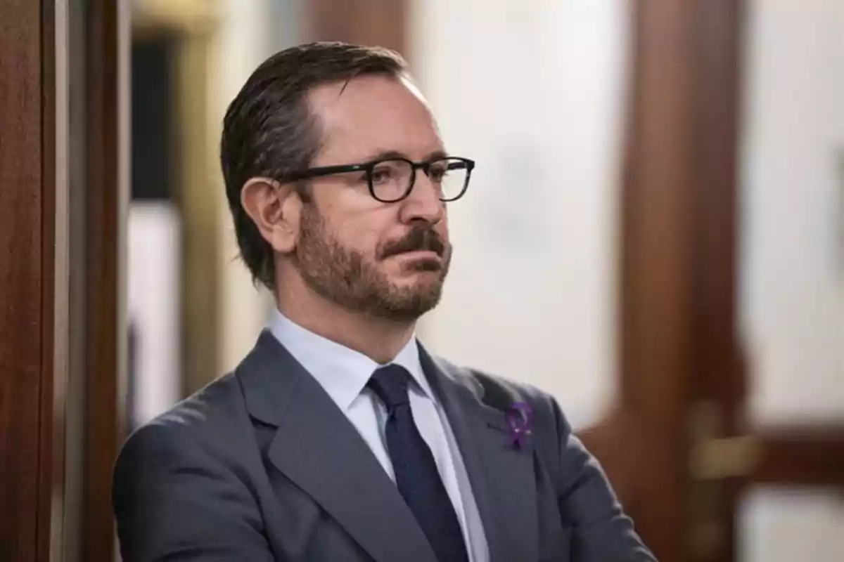 Hombre de traje gris con gafas y barba de pie junto a una pared en un entorno interior Hombre de traje gris con gafas y barba de pie junto a una pared en un entorno interior