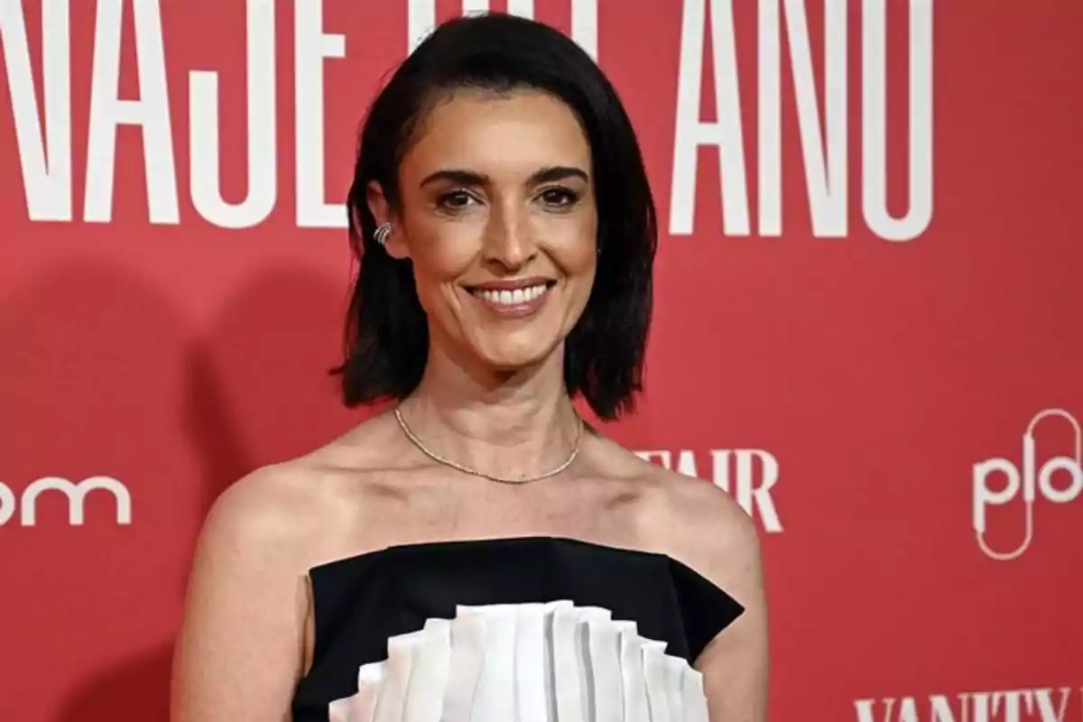 Mujer de cabello oscuro y corto sonriendo frente a un fondo rojo con letras blancas, vestida con un vestido negro y blanco sin tirantes