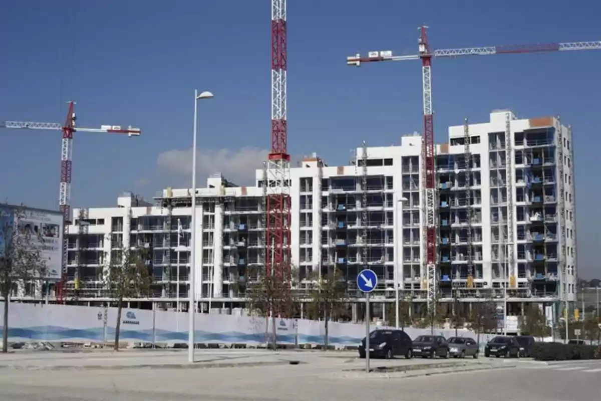 Edificio de departamentos en construcción con varias grúas y autos estacionados al frente en un día soleado