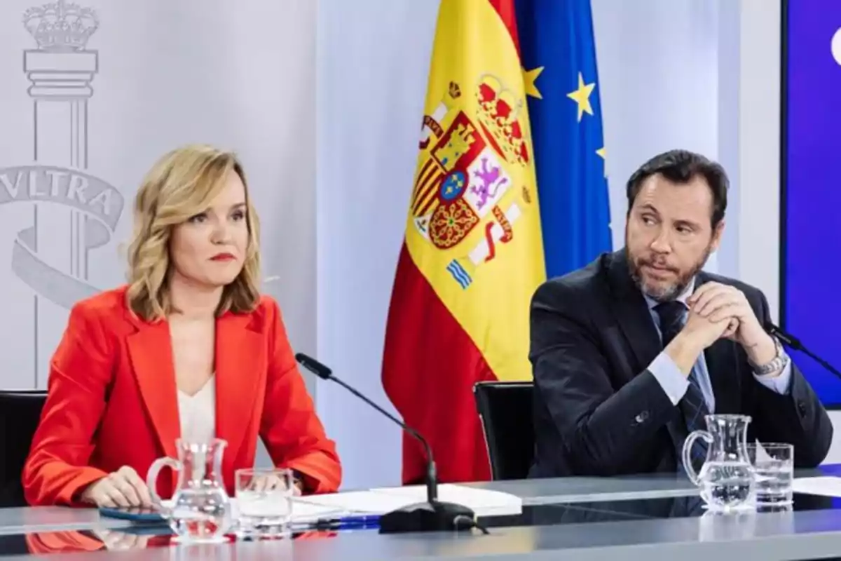 Dos personas sentadas en una mesa durante una conferencia de prensa con banderas de España y la Unión Europea al fondo Dos personas sentadas en una mesa durante una conferencia de prensa con banderas de España y la Unión Europea al fondo
