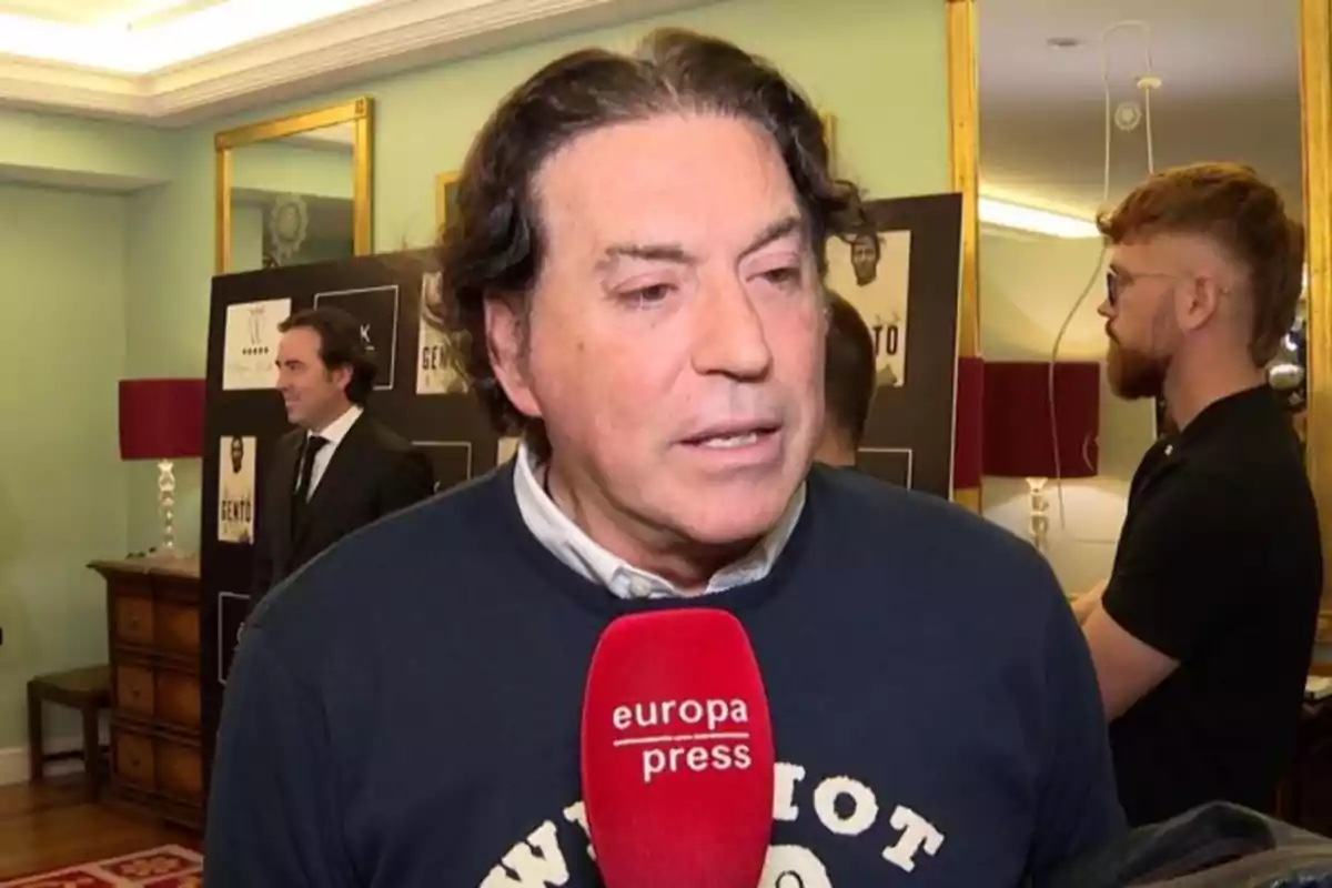 Un hombre de cabello oscuro sostiene un micrófono rojo de europa press mientras es entrevistado en un evento con varias personas y carteles en el fondo.