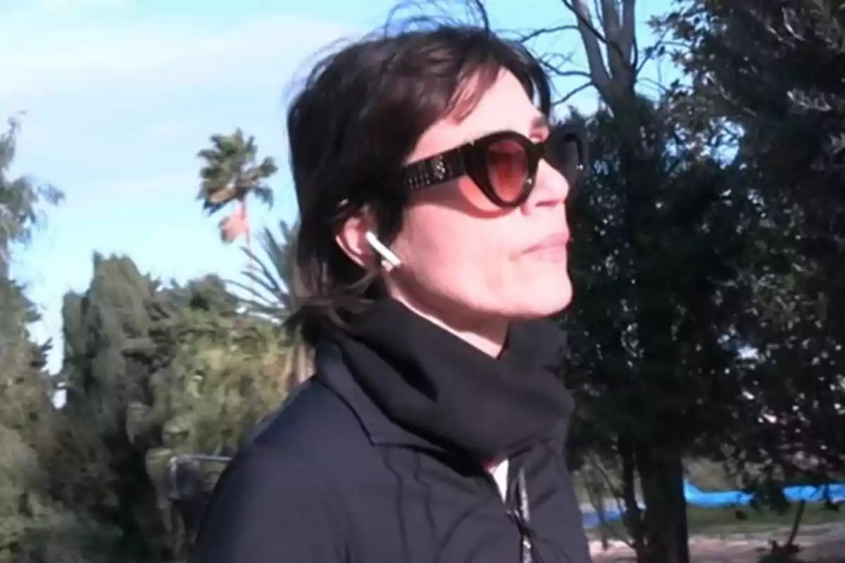 Mujer con gafas de sol y auriculares caminando al aire libre rodeada de árboles Mujer con gafas de sol y auriculares caminando al aire libre rodeada de árboles