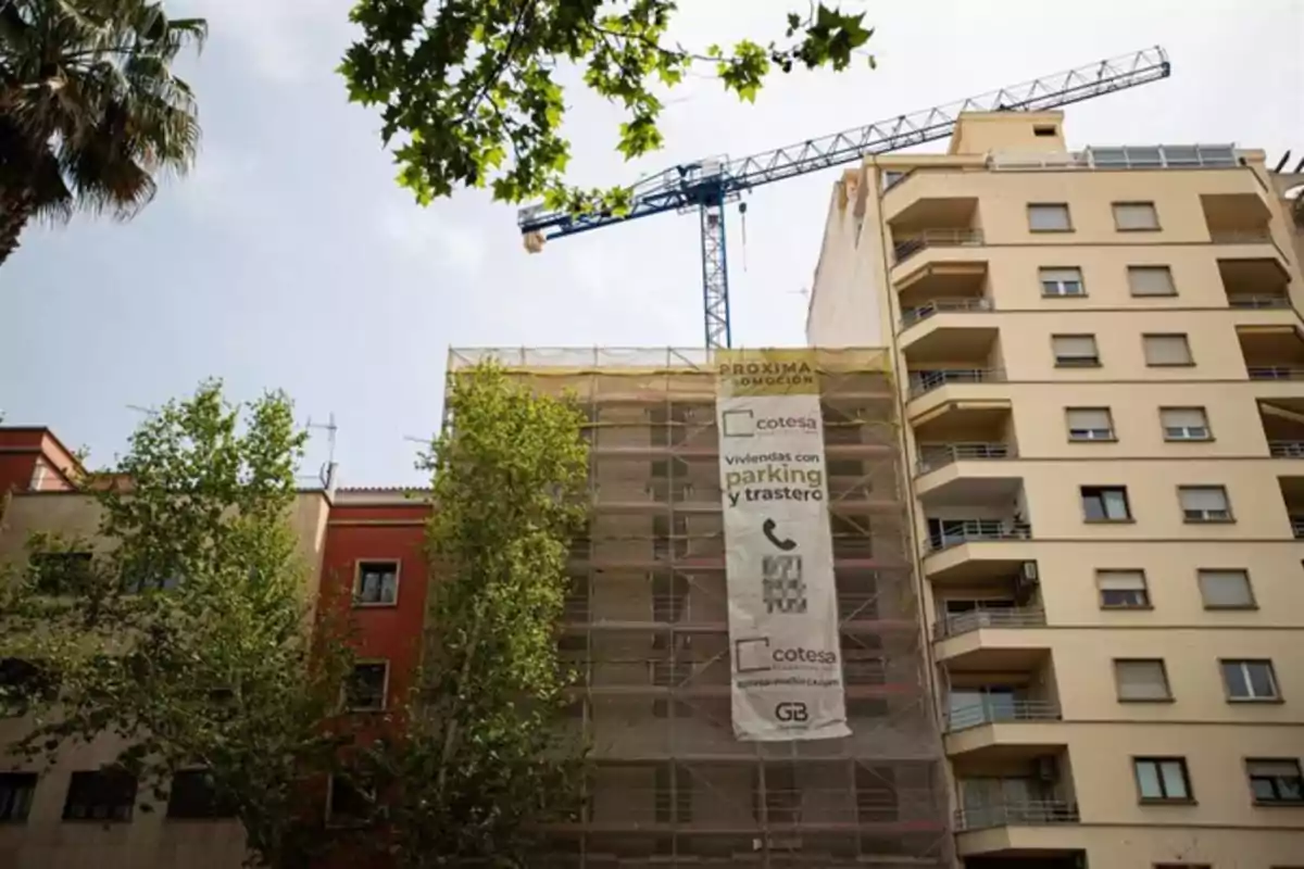 Edificio en construcción cubierto con andamios y una lona publicitaria, junto a otros edificios residenciales y árboles, con una grúa azul al fondo Edificio en construcción cubierto con andamios y una lona publicitaria, junto a otros edificios residenciales y árboles, con una grúa azul al fondo