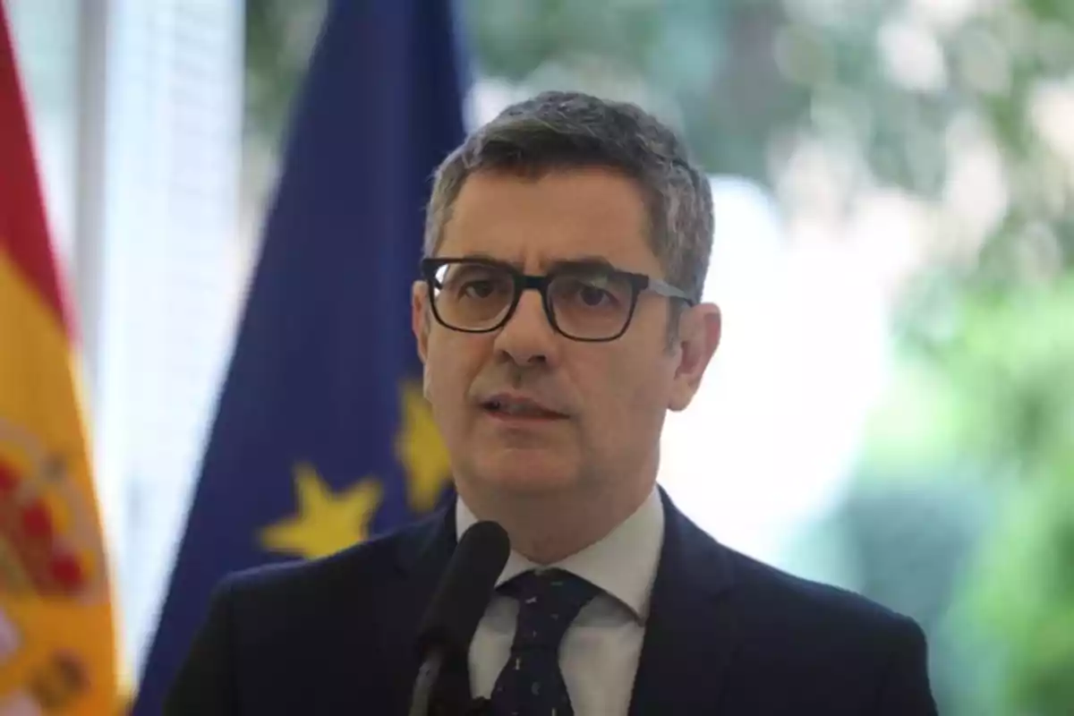 Hombre de cabello canoso y gafas hablando frente a un micrófono con banderas de España y la Unión Europea al fondo Hombre de cabello canoso y gafas hablando frente a un micrófono con banderas de España y la Unión Europea al fondo