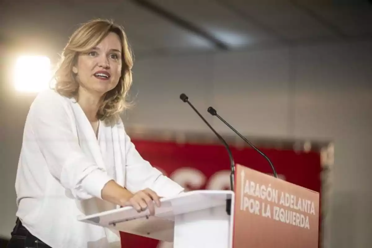 Mujer de cabello rubio y ropa blanca hablando en un atril con micrófonos en un evento político donde se lee Aragón adelanta por la izquierda Mujer de cabello rubio y ropa blanca hablando en un atril con micrófonos en un evento político donde se lee Aragón adelanta por la izquierda