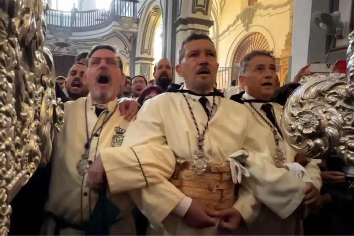 Hombres vestidos con túnicas blancas y medallas participan en una procesión religiosa dentro de una iglesia, mostrando expresiones de emoción y esfuerzo mientras cargan un paso decorado. Hombres vestidos con túnicas blancas y medallas participan en una procesión religiosa dentro de una iglesia, mostrando expresiones de emoción y esfuerzo mientras cargan un paso decorado.
