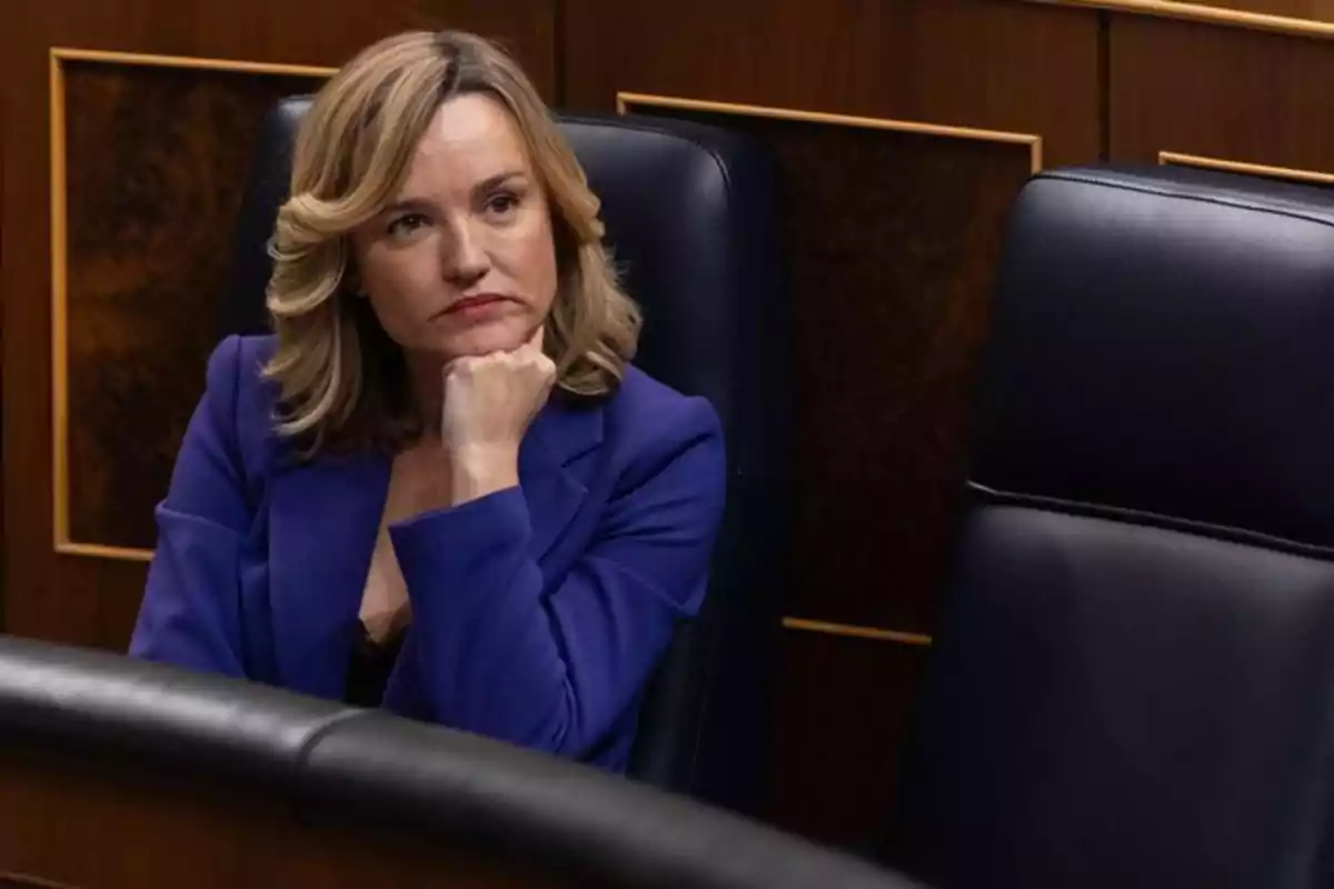 Mujer rubia con chaqueta azul sentada en una silla de un recinto oficial con expresión pensativa y la mano apoyada en el mentón Mujer rubia con chaqueta azul sentada en una silla de un recinto oficial con expresión pensativa y la mano apoyada en el mentón