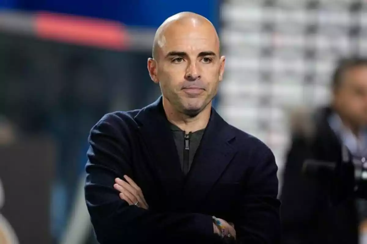 Hombre calvo con chaqueta oscura y brazos cruzados en un entorno deportivo Hombre calvo con chaqueta oscura y brazos cruzados en un entorno deportivo