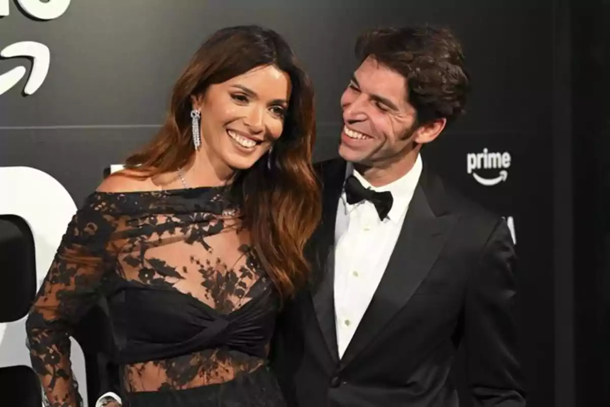 Una pareja sonríe elegantemente vestida en un evento de gala con fondo negro y logotipos de Prime.