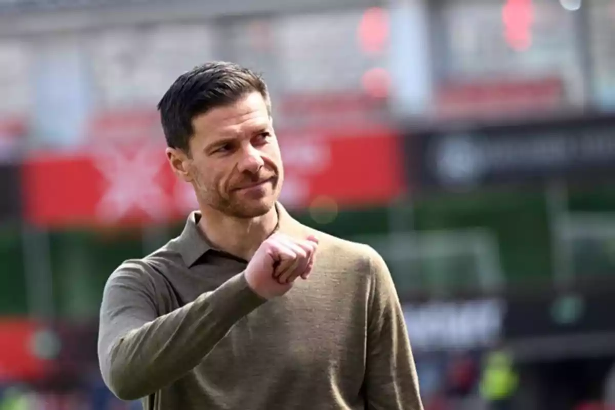Hombre con barba corta y cabello oscuro vestido con suéter marrón claro haciendo un gesto con la mano en un estadio desenfocado Hombre con barba corta y cabello oscuro vestido con suéter marrón claro haciendo un gesto con la mano en un estadio desenfocado