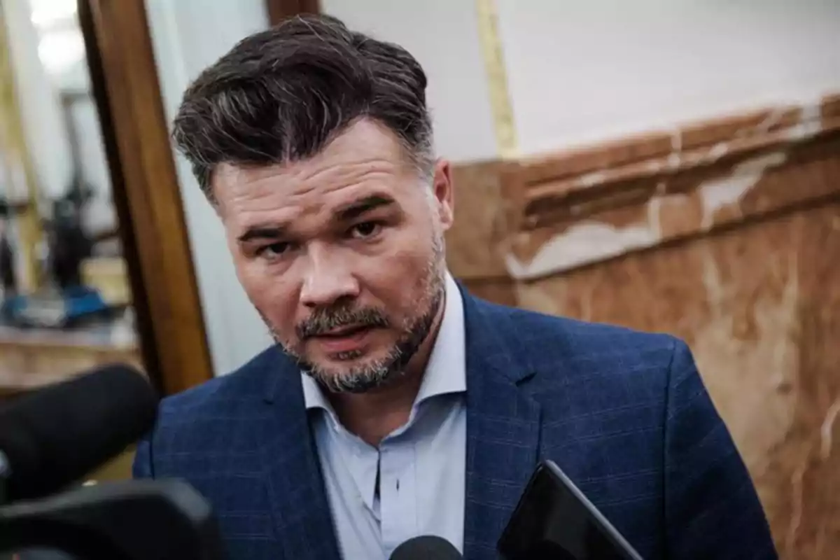 Hombre de cabello oscuro y barba con saco azul hablando frente a varios micrófonos en un interior elegante Hombre de cabello oscuro y barba con saco azul hablando frente a varios micrófonos en un interior elegante