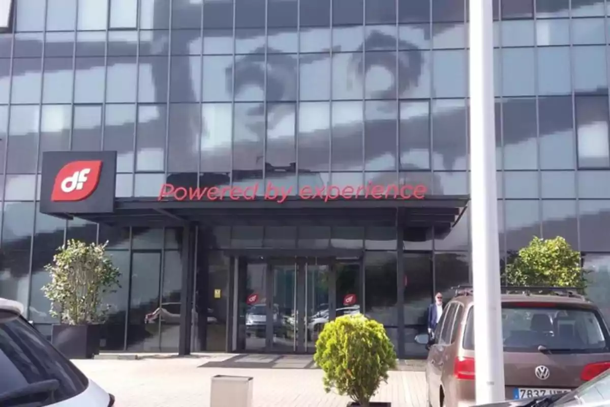 Fachada de edificio de oficinas con letrero rojo que dice Powered by experience y logotipo df a la izquierda, con autos estacionados y plantas al frente Fachada de edificio de oficinas con letrero rojo que dice Powered by experience y logotipo df a la izquierda, con autos estacionados y plantas al frente