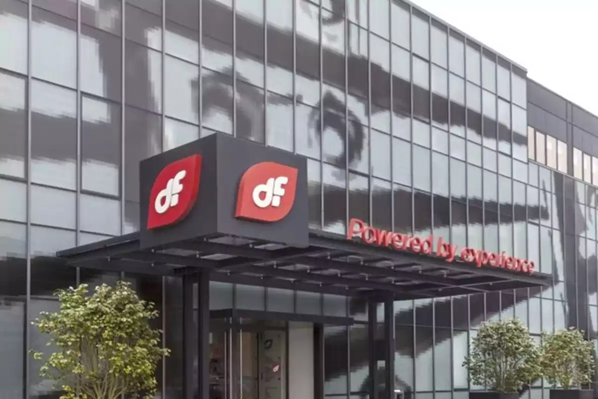 Fachada de edificio moderno con logotipo rojo y blanco de la empresa DF y el lema Powered by experience junto a la entrada principal rodeada de plantas Fachada de edificio moderno con logotipo rojo y blanco de la empresa DF y el lema Powered by experience junto a la entrada principal rodeada de plantas