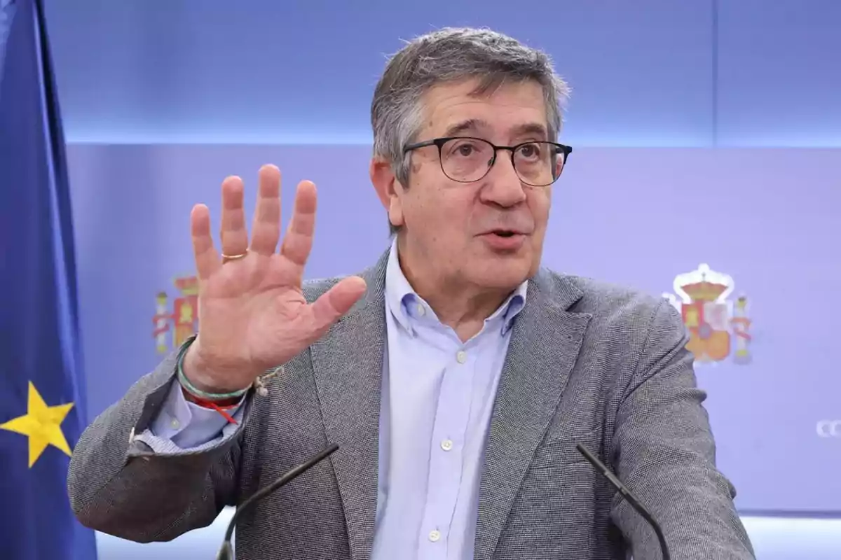 Hombre de cabello canoso y gafas hablando en una rueda de prensa con la mano levantada y una bandera de la Unión Europea al fondo Hombre de cabello canoso y gafas hablando en una rueda de prensa con la mano levantada y una bandera de la Unión Europea al fondo