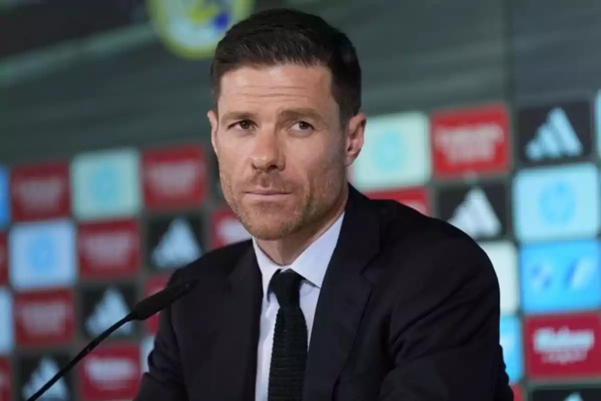 Hombre con traje oscuro y corbata negra sentado frente a un micrófono en una conferencia de prensa con fondo de logotipos deportivos desenfocados